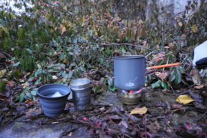 Picture of a small alcohol stove with a small pot on it heating water for tea with two cups, one a silicone collapsible backpacking cup and one a pewter tumbler.